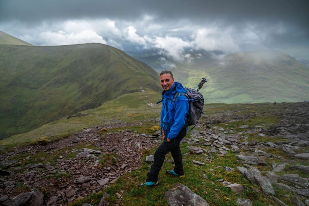 Founder of WildnHappytravel.com stands on the crest of the highest mountain in Ireland called Carrauntoohil. Hiking in Kerry. The weather is foggy, he is wearing a blue waterproof jacket and carrying a backpack with trekking poles