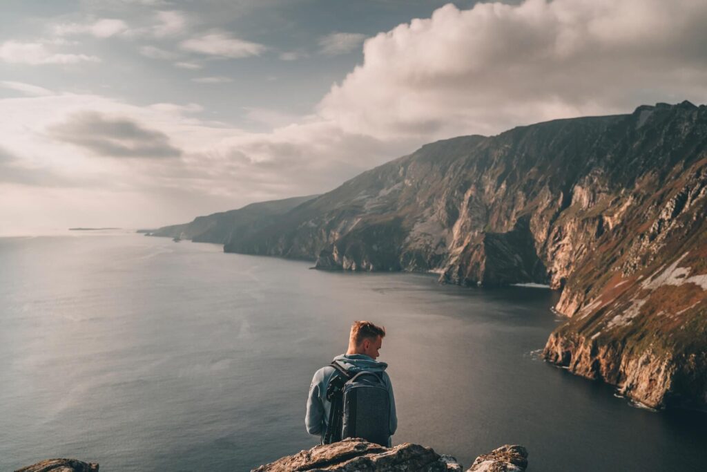 A man standing on top of a mountain overlooking a large body of water on a cloudy day. Photo: Lukas Medvedevas. Killarney
