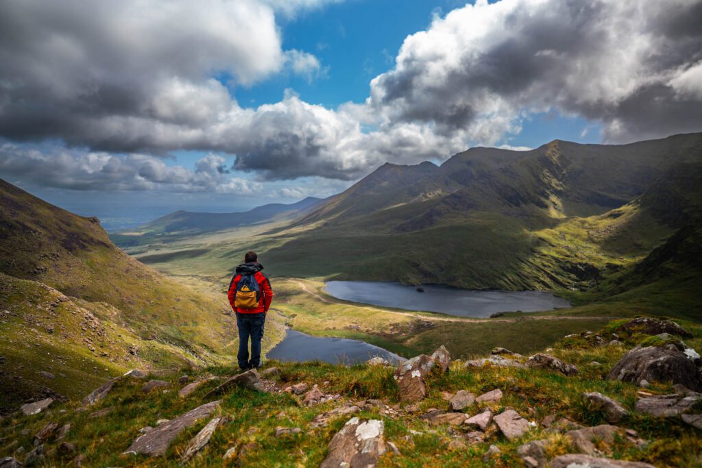 a man in a black-and-red outdoor jacket and a backpack stands at a viewpoint in the valley. There are two glacial lakes in the valley. Ireland's highest mountains spread out around. It's a rainy summer day